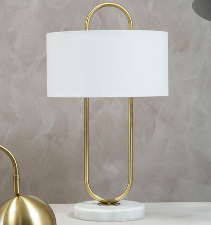 Table lamp with gold base and white shade on a marble surface with a gray textured wall background