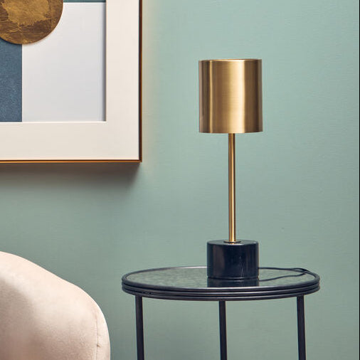 Beige armchair with a gray pillow, gold-framed abstract art, and a small round table with a lamp against a green wall.