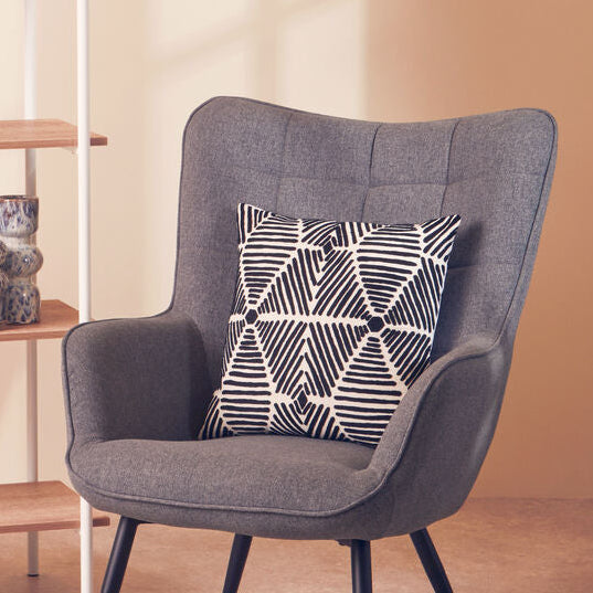 Gray armchair with a patterned pillow next to a wooden shelf against a beige wall.
