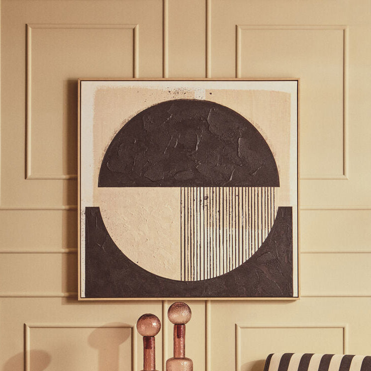 A styled interior scene with a beige paneled wall, a framed abstract artwork above a striped armchair, a ribbed round coffee table, and decorative glass bottles on a side table.