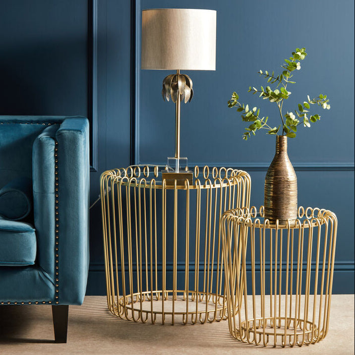 Gold wire side tables with a lamp and plant against a blue wall.