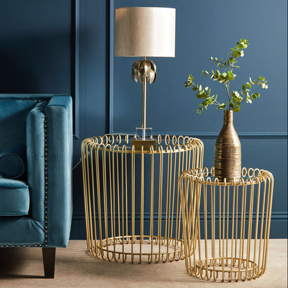 Gold wire side tables with a lamp and plant against a blue wall.