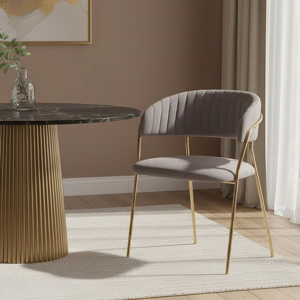 Modern dining table with marble top and gold base, accompanied by a gray chair with gold legs in a room with brown walls and white curtains.