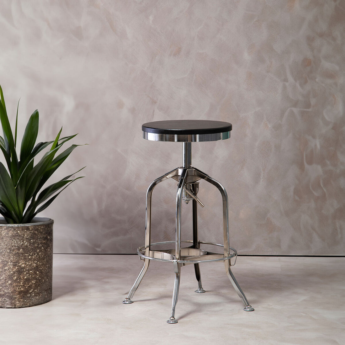 Metal bar stool with black seat next to a potted plant against a textured wall.