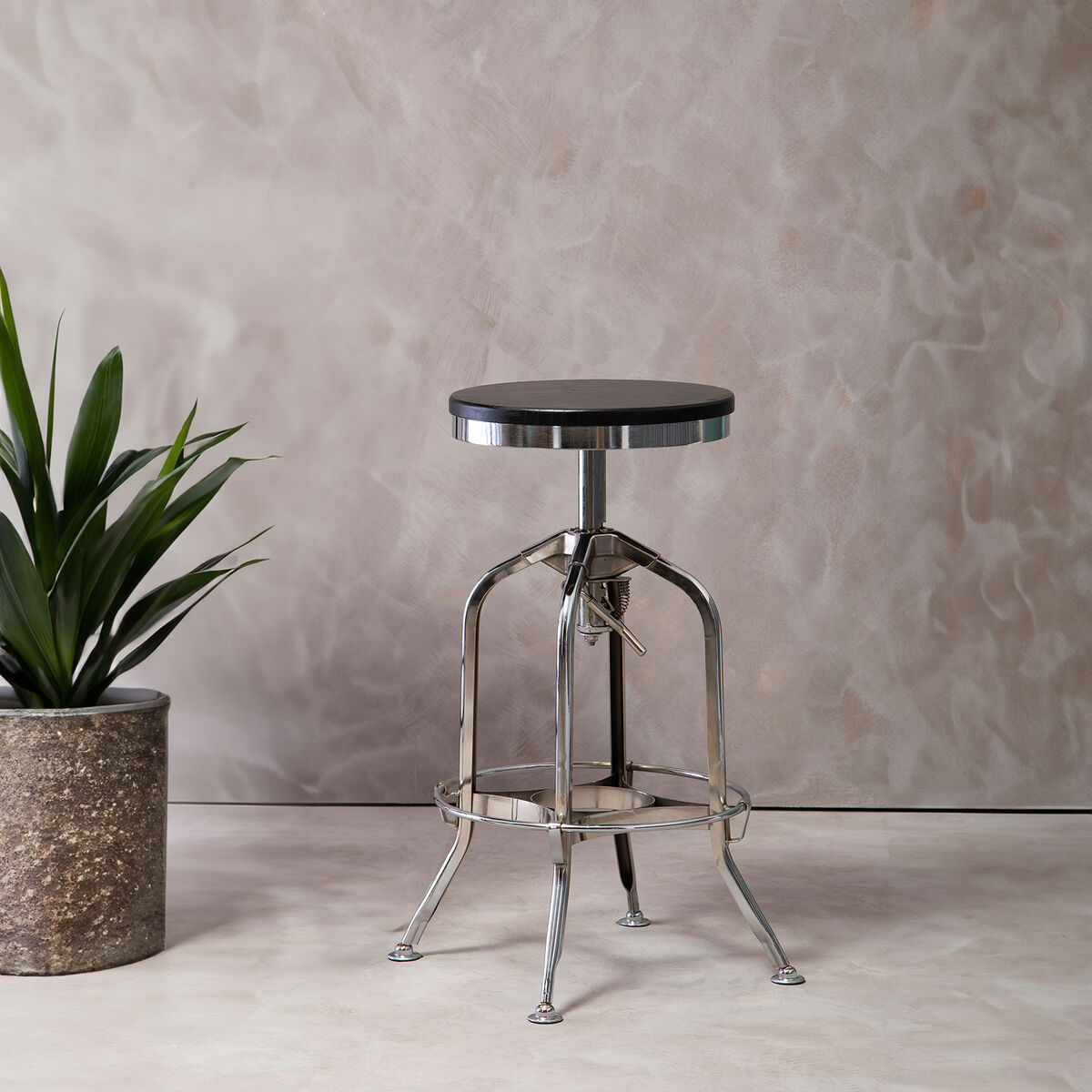 Metal bar stool with black seat next to a potted plant against a textured wall.