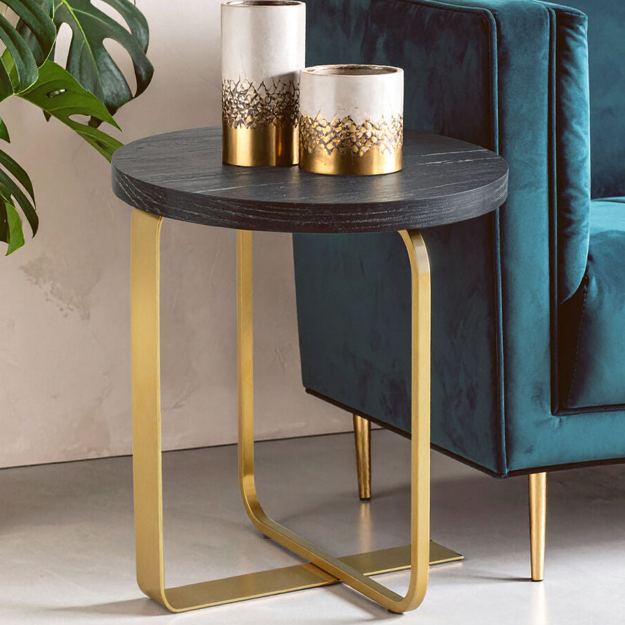 Teal sofa with a small round table featuring gold legs and two decorative items, with a plant in the corner.