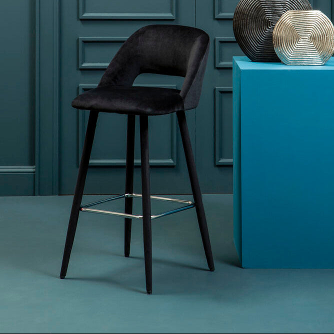Dark brown bar stool in front of a blue pedestal with decorative items against a gray wall.