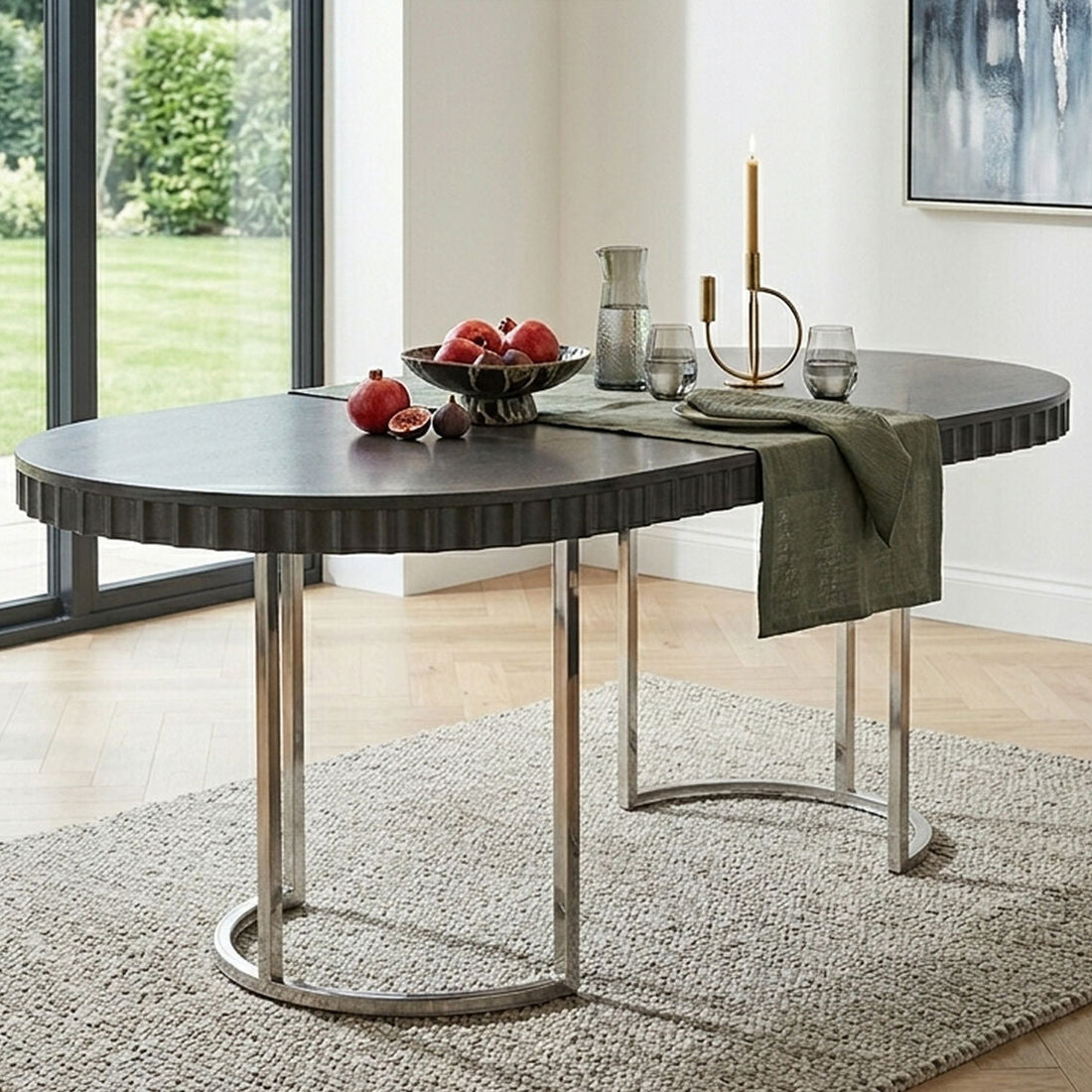An oval dining table with a dark gray ribbed top and polished silver metal legs, styled in a bright modern dining room with a green throw blanket, fruit bowl, and glassware.
