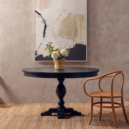 Round black table with a vase of flowers and a wooden chair against a textured wall with abstract art.