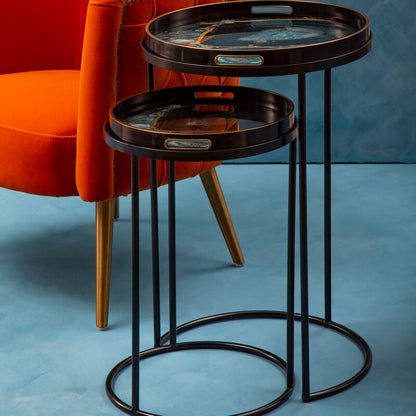 Two round wooden trays on a black metal stand next to an orange chair against a blue textured wall.