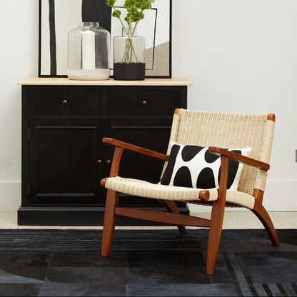Wicker chair with wooden armrests and a black and white cushion in front of a black cabinet.