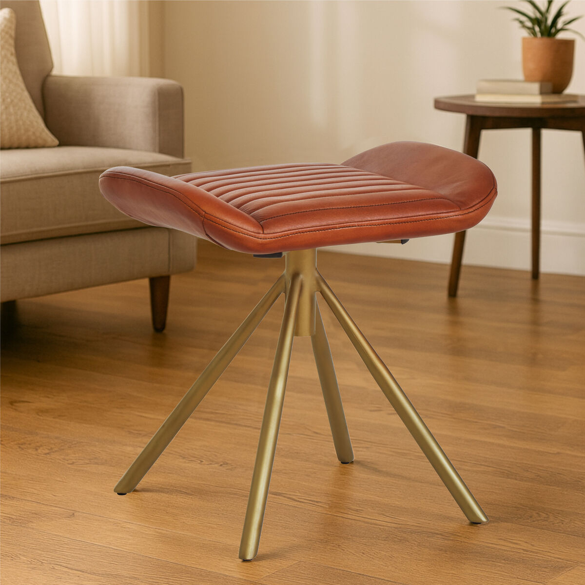 Brown leather stool with gold legs on a wooden floor in a living room setting.