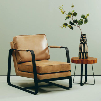 Brown leather armchair with black metal frame next to a wooden side table with a vase on a light green background