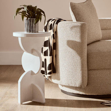 Beige armchair with a patterned blanket and a small white side table with a plant in a living room.