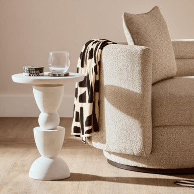 Beige armchair with a patterned throw blanket next to a white side table in a living room setting.