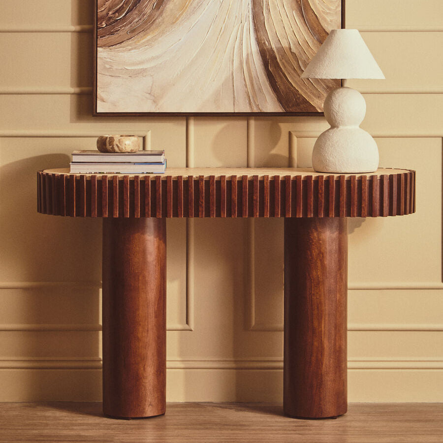 A modern console table with a fluted wood top and two chunky cylindrical legs in warm medium brown tones, styled in an interior setting with a white sculptural table lamp, books, and a large abstract painting on a beige paneled wall.