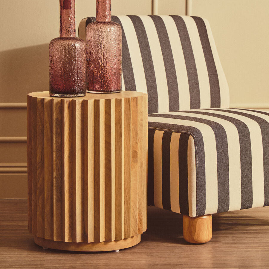 A modern cylindrical wooden side table with vertical fluting, holding two textured rose-gold glass bottle vases, set next to a black and cream striped armchair in a warm interior room.