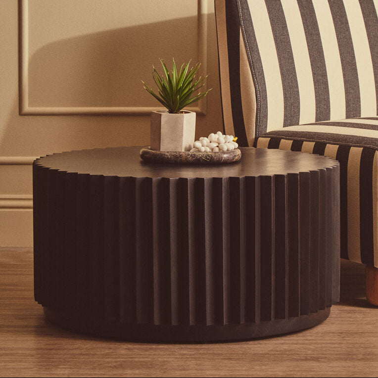 A dark brown fluted round coffee table in a styled living room setting next to a cream and dark brown striped armchair.