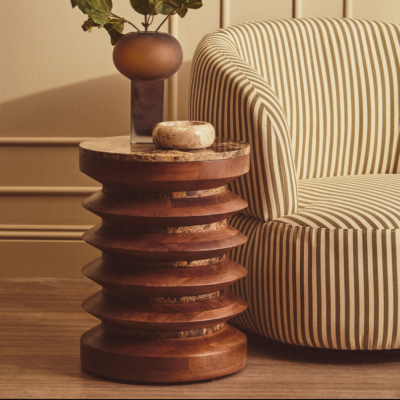 A styled interior scene showing a round upholstered armchair with beige and brown vertical stripes next to a sculptural wooden side table topped with a dark vase of greenery and a small bowl.