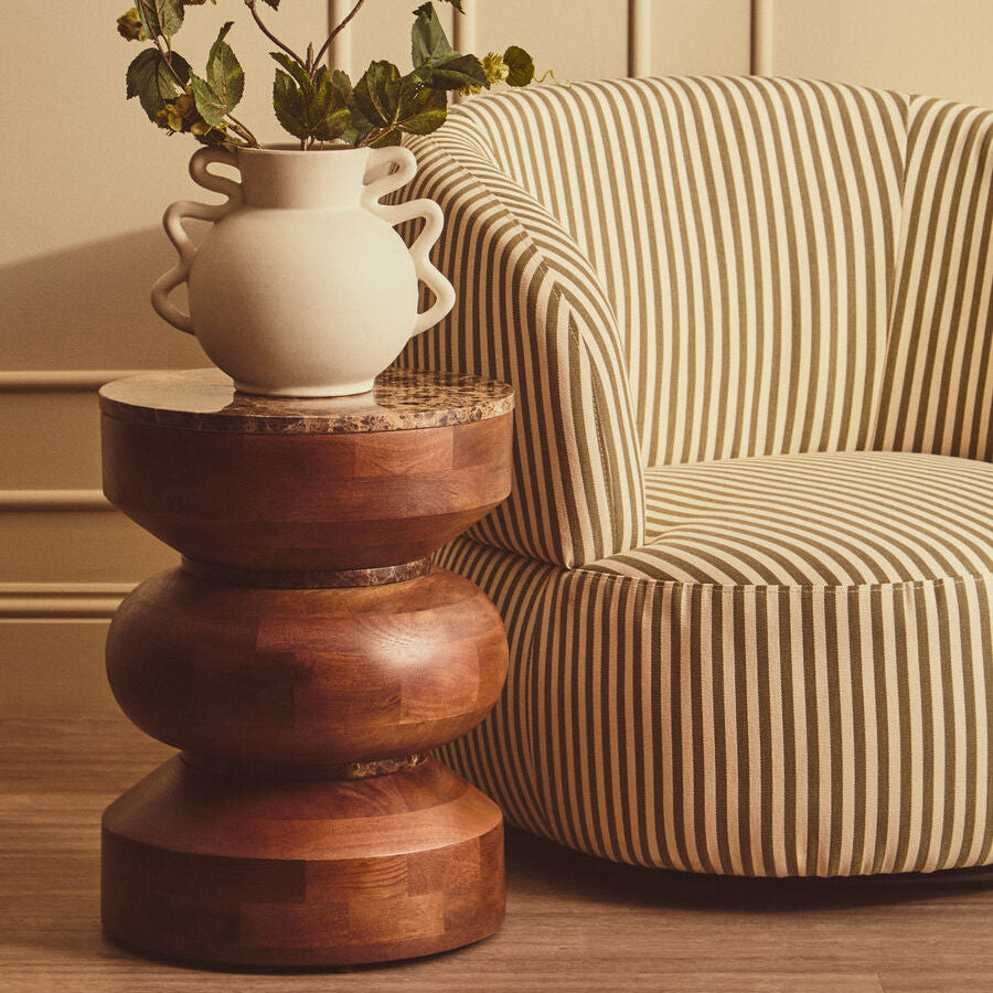 A rustic wooden tiered side table holding a cream ceramic vase with green leaves, positioned next to a beige and brown striped armchair in a warm, neutral-toned room.