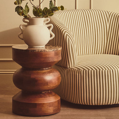 A rustic wooden tiered side table holding a cream ceramic vase with green leaves, positioned next to a beige and brown striped armchair in a warm, neutral-toned room.
