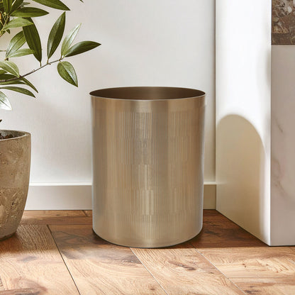 Bronze-colored cylindrical trash can on a wooden floor with a plant in the background