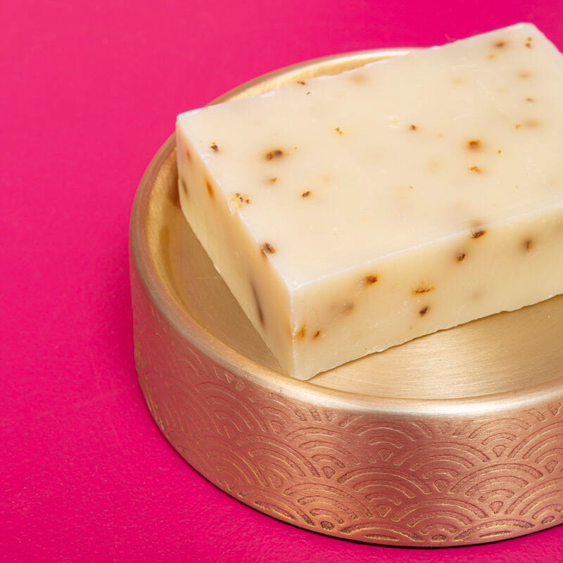 Two pieces of white soap with brown specks on a gold soap dish against a pink background