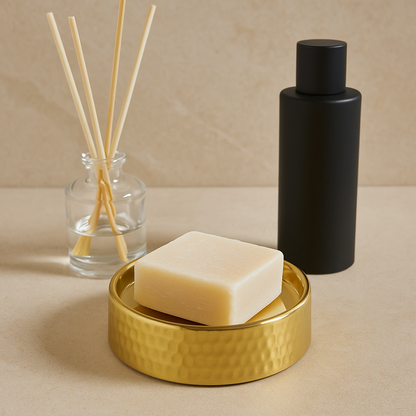 Bar of soap on a gold dish with a black bottle and reed diffuser in the background.