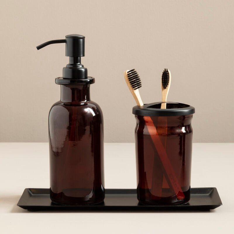 Brown glass soap dispenser and toothbrush holder on a black tray with a beige background