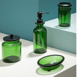 Set of green glass bathroom accessories including a bottle, jar, and soap dish on a white surface with a teal wall.