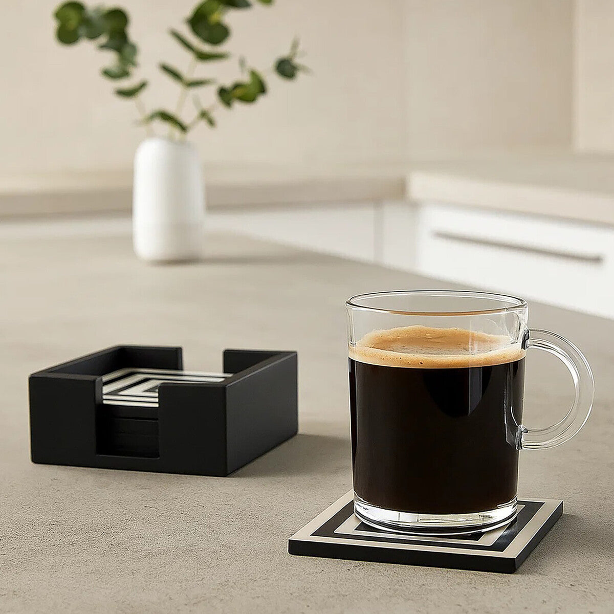 Clear glass mug with coffee on a coaster next to a black coaster holder on a light surface.