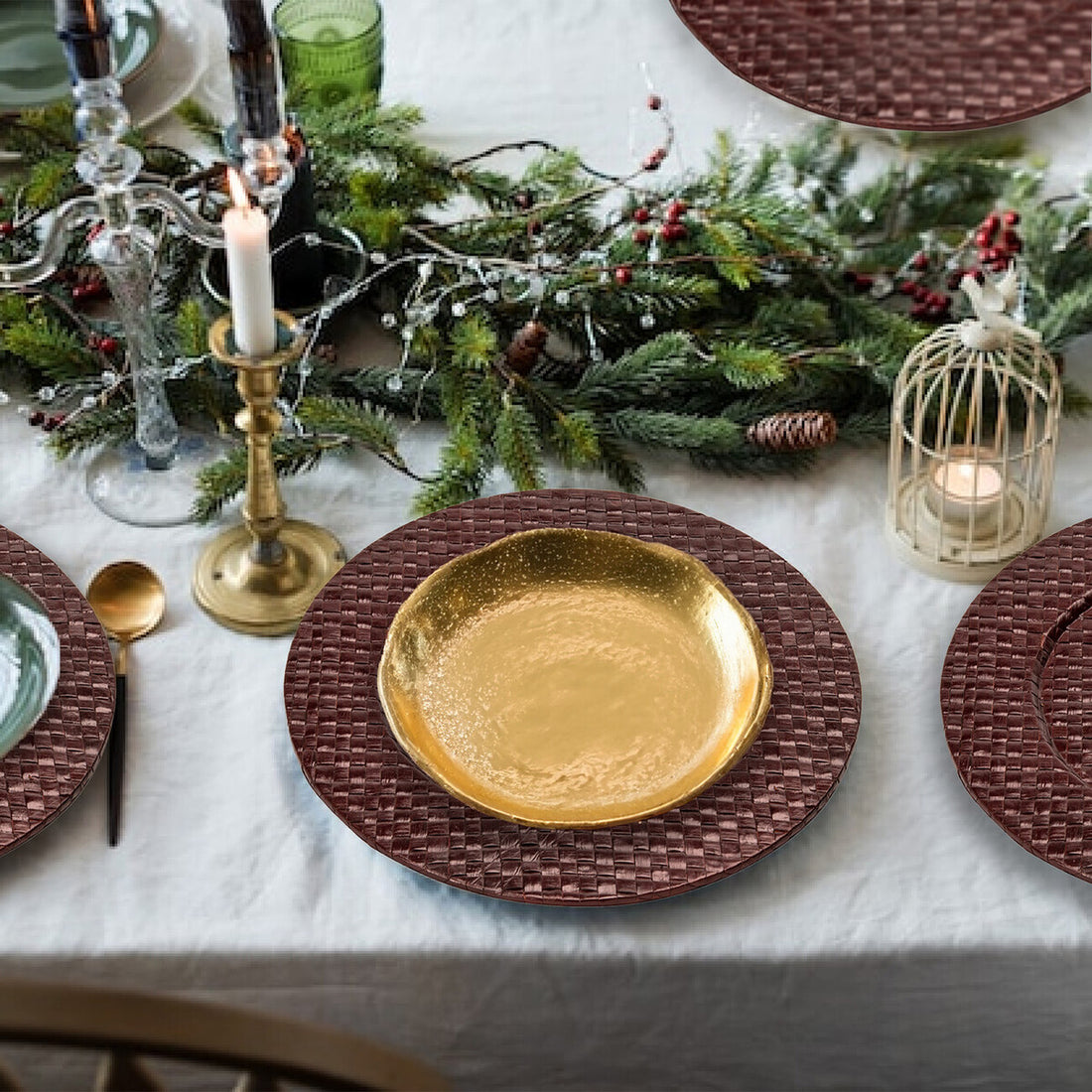 Decorative table setting with gold plate, candles, and greenery on a white tablecloth.