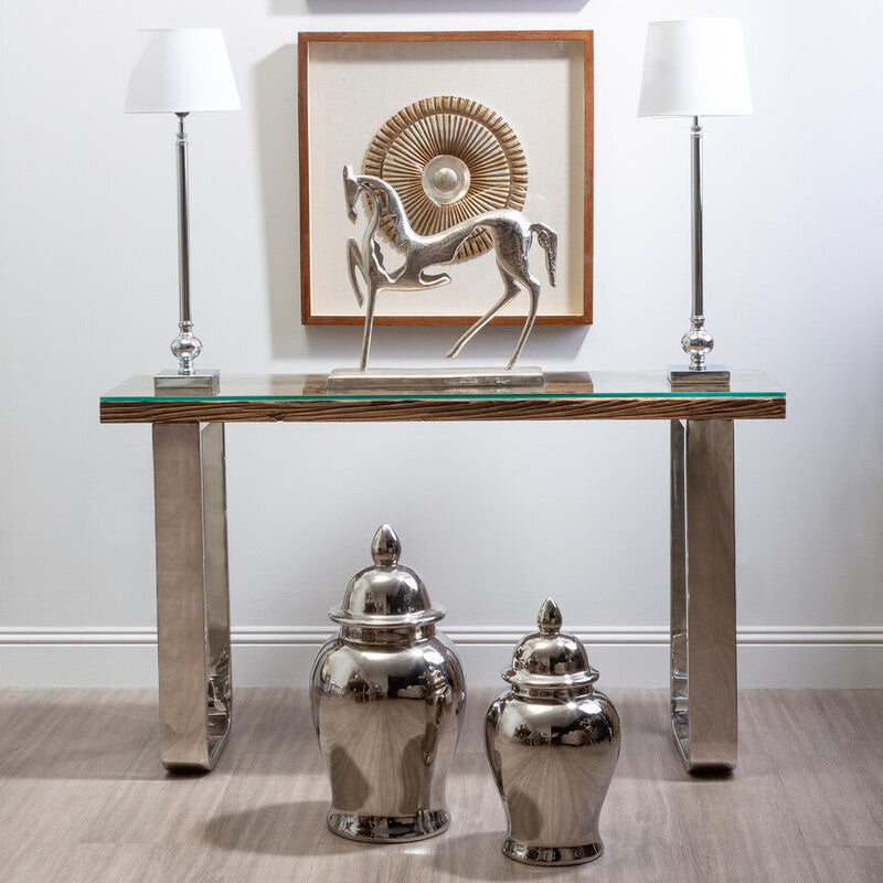 Decorative console table with silver urns, a glass top, and framed artwork on a white wall.