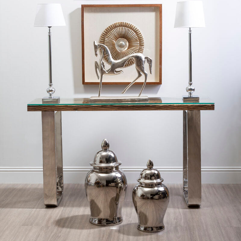 Decorative console table with silver vases, a glass top, and framed artwork on a white wall.