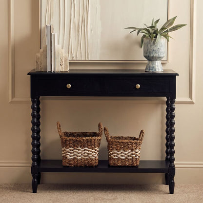 A black wooden console table with a single drawer, two brass knobs, and a lower shelf holding two woven baskets, styled in a warm beige room under a mirror.