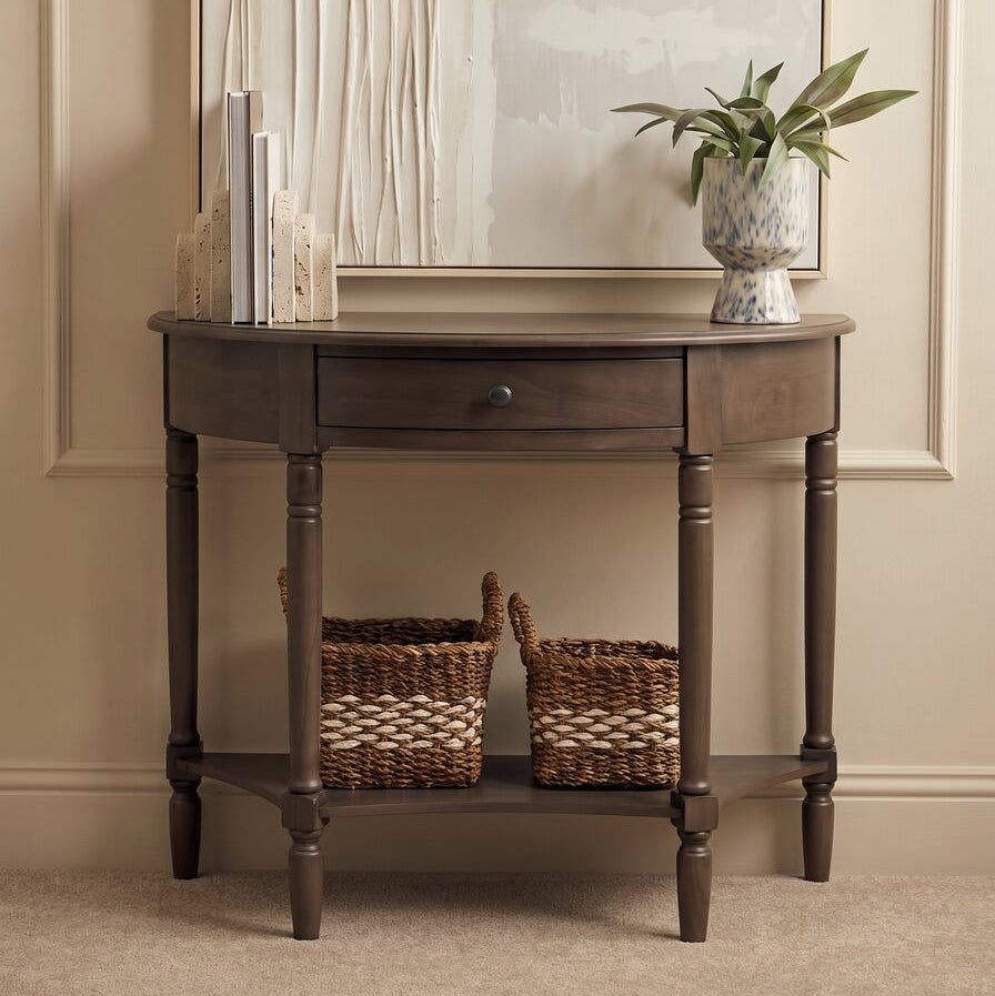 A dark brown half-console table with a single drawer and open lower shelf, styled in a home entryway under a mirror with a potted plant and baskets on top.