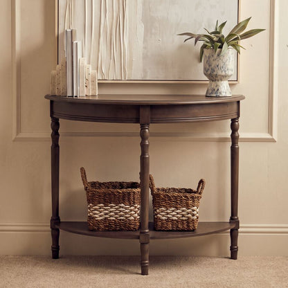A dark brown wooden console table with a half-round shape, featuring turned legs, a lower shelf with two woven storage baskets, and a tabletop styled with books and a potted plant, set against a beige paneled wall.