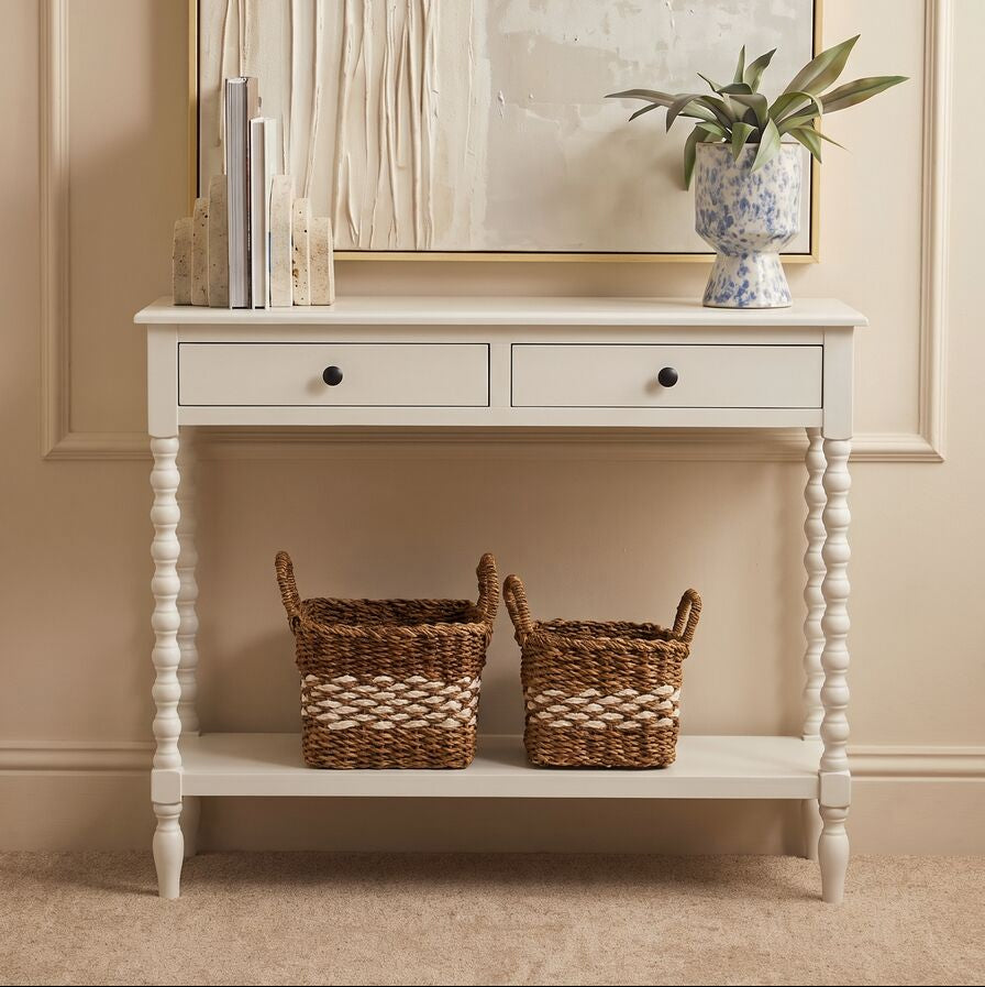 A white wooden console table with two drawers and a lower shelf, styled in a warm beige room with two woven baskets, books, and a potted plant.