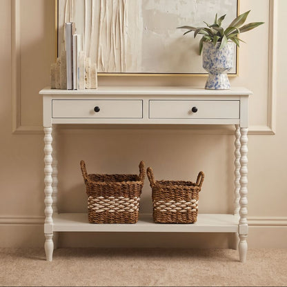 A white wooden console table with two drawers and a lower shelf, styled in a warm beige room with two woven baskets, books, and a potted plant.