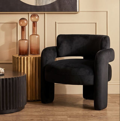 A styled interior scene with a black velvet armchair, a dark ribbed coffee table, decorative amber bottles on a gold side table, and a large abstract wall art piece above a beige paneled wall.