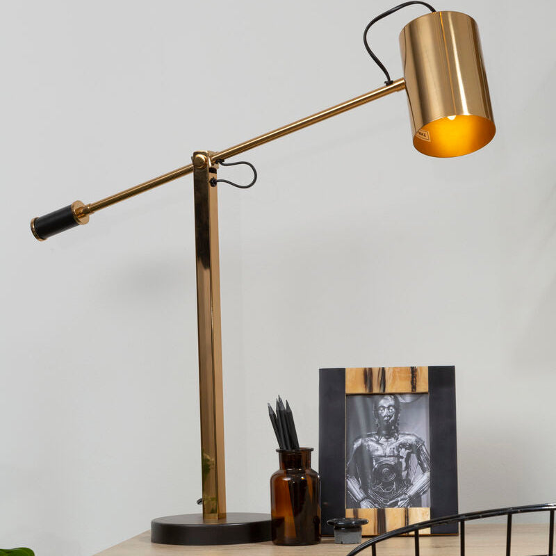 Gold desk lamp on a wooden desk with a black chair and framed picture in the background.