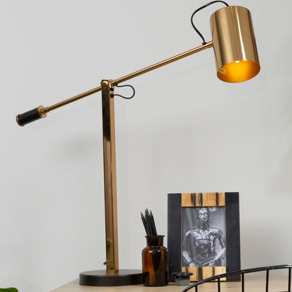 Gold desk lamp on a wooden desk with a black chair and framed picture in the background.